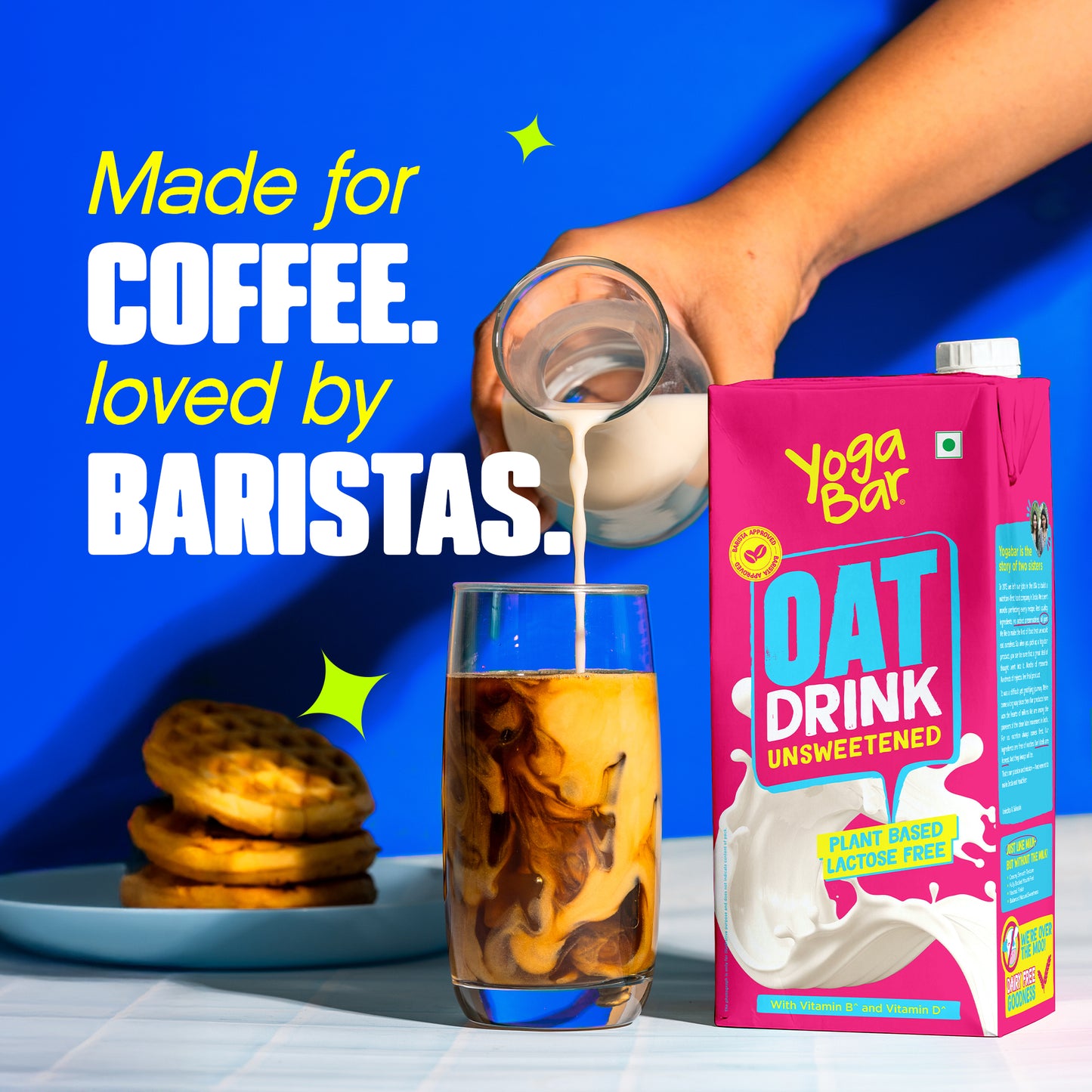 Yoga Bar Unsweetened Oat Milk being poured into iced coffee next to a plate of waffles on a blue background.